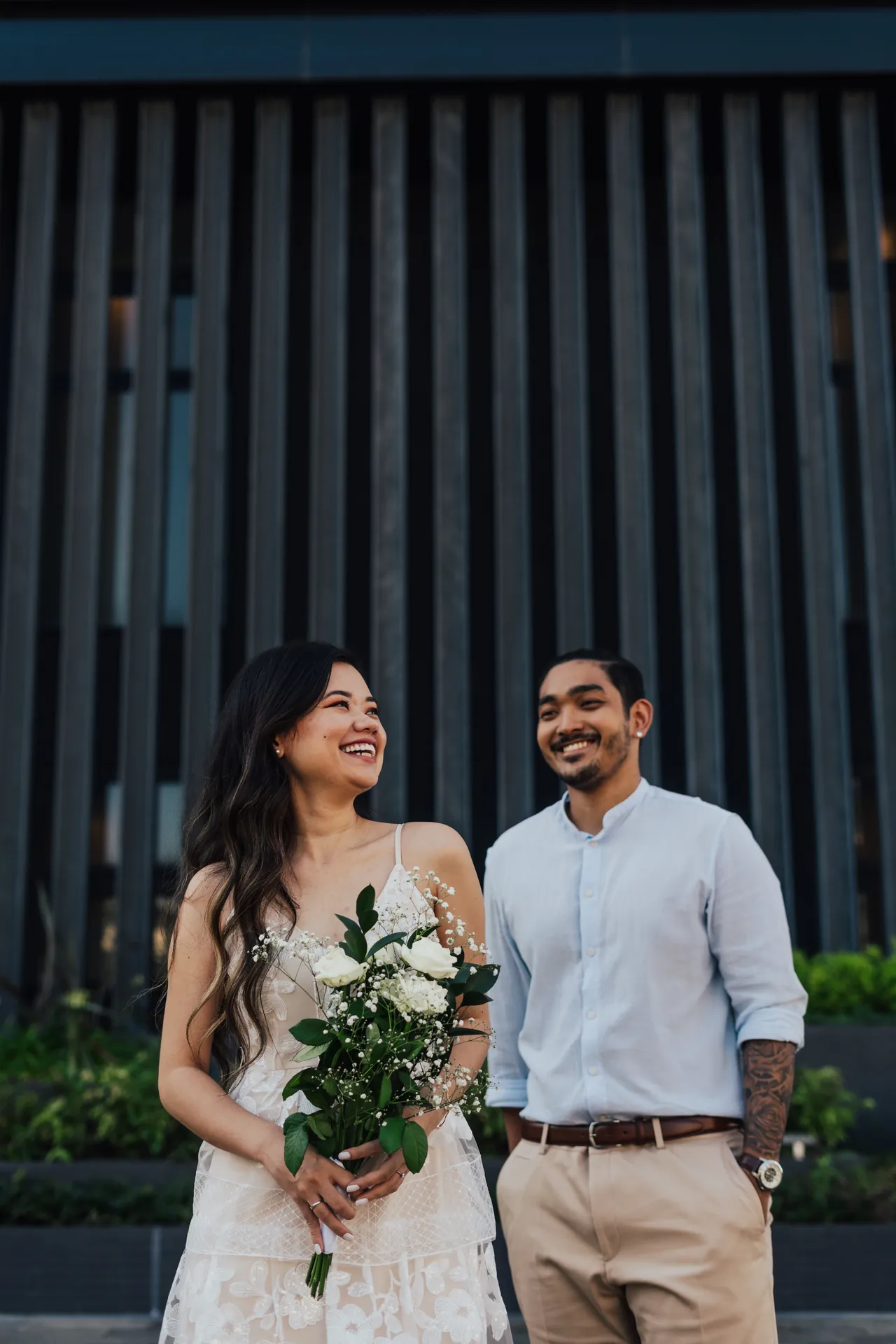 Casal sorrindo lado a lado, ela com um buquê de flores brancas
